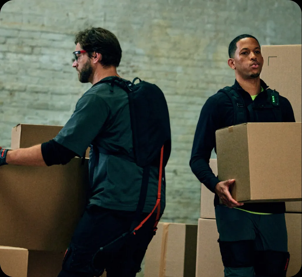 Two warehouse workers wearing SafeLift exosuits carrying cardboard boxes